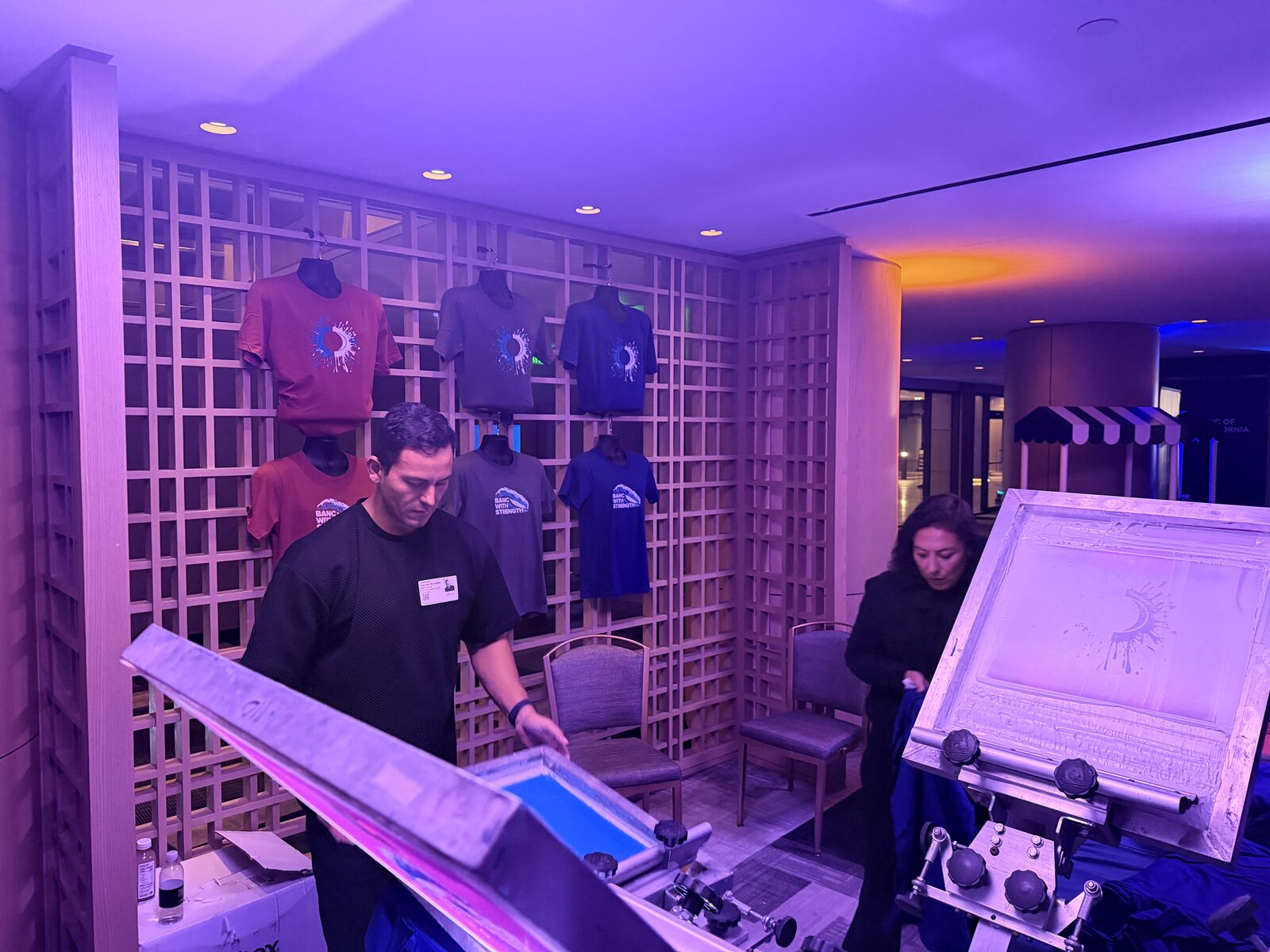 Wall of custom event shirts behind a live screen printing station