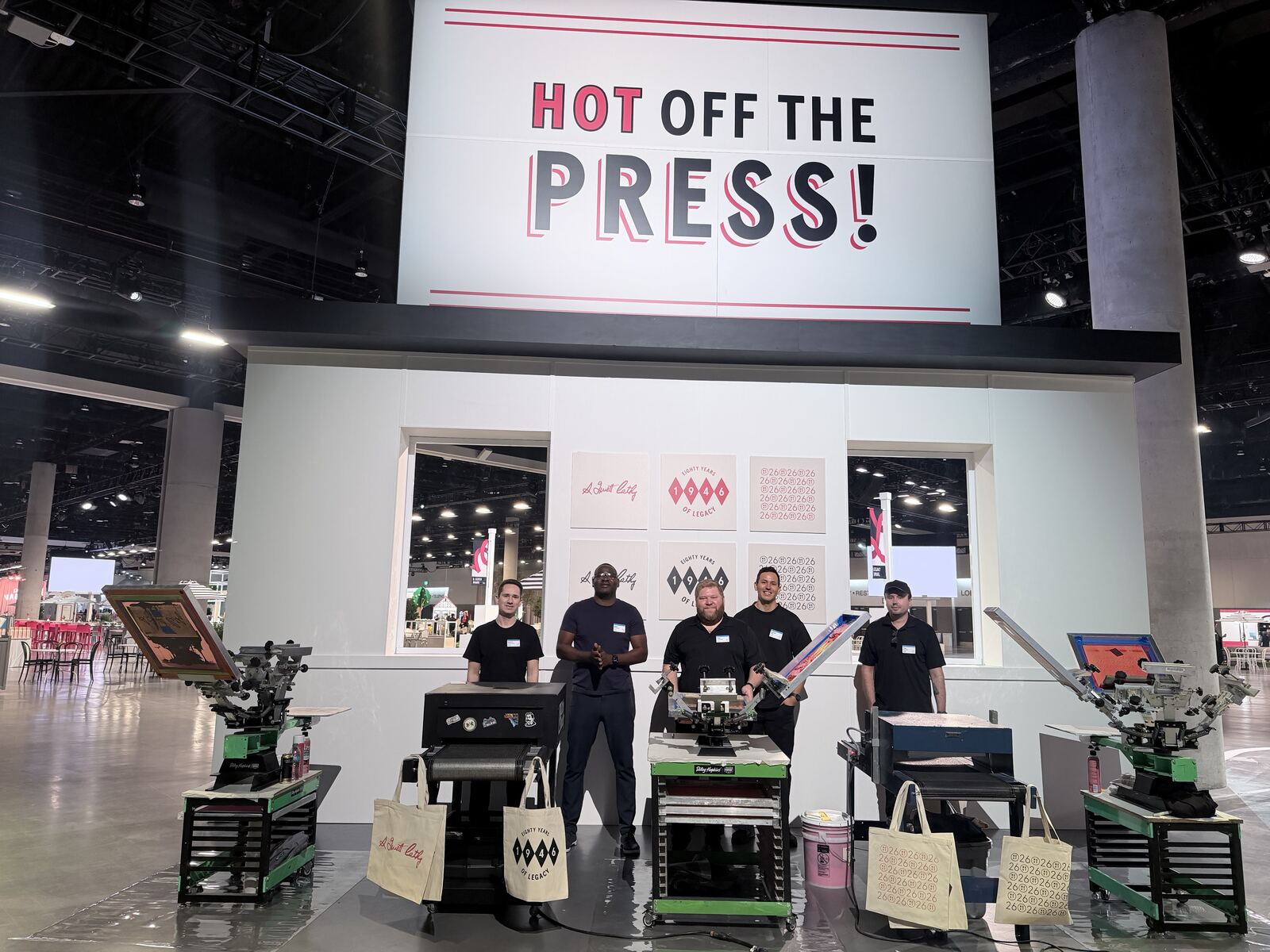 Professional trade show booth with screen-printed totes and t-shirts displayed on a white stand as staff present to visitors